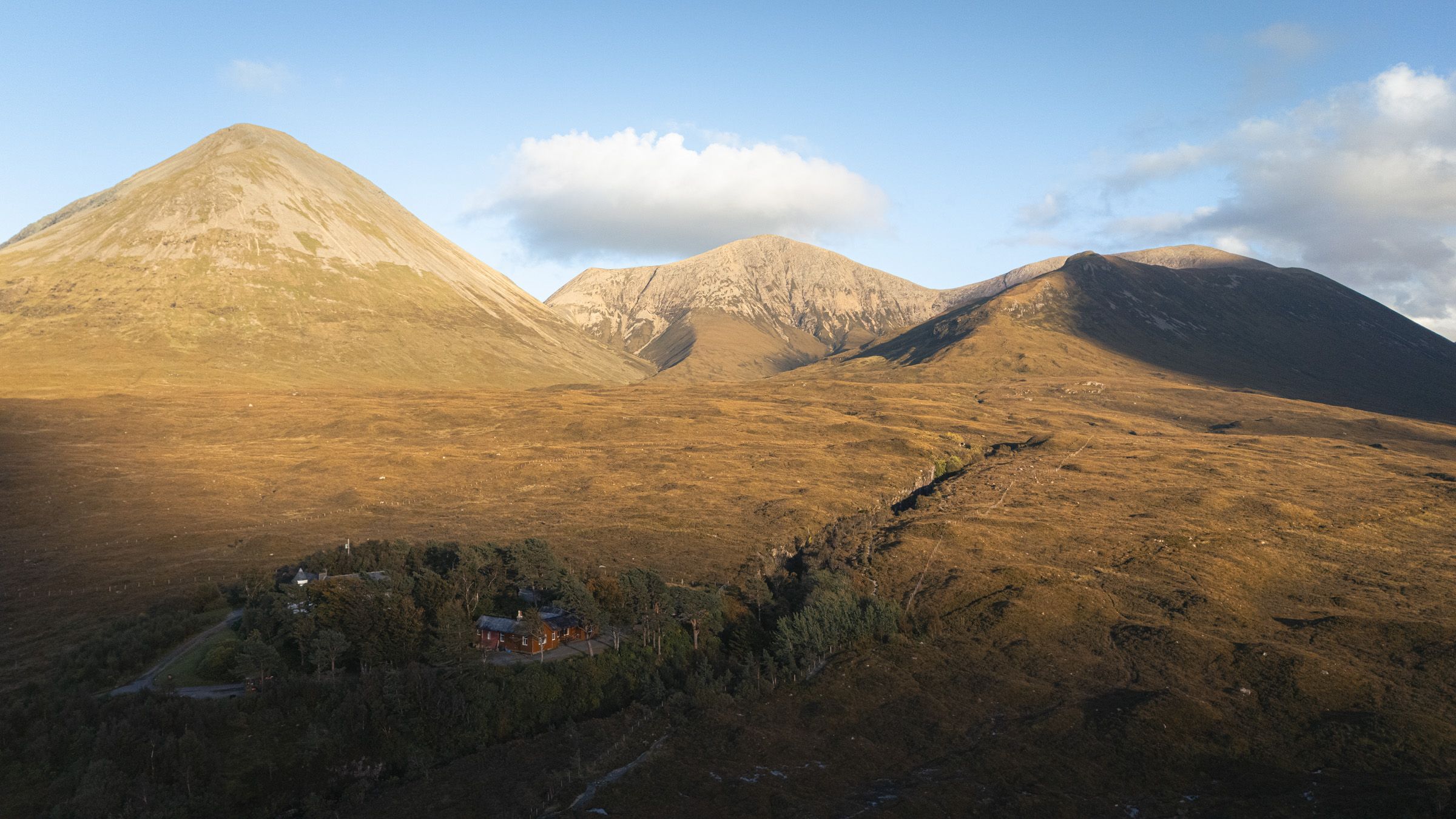 Aerial view of a small house among trees on a golden moor, below three sunlit mountains