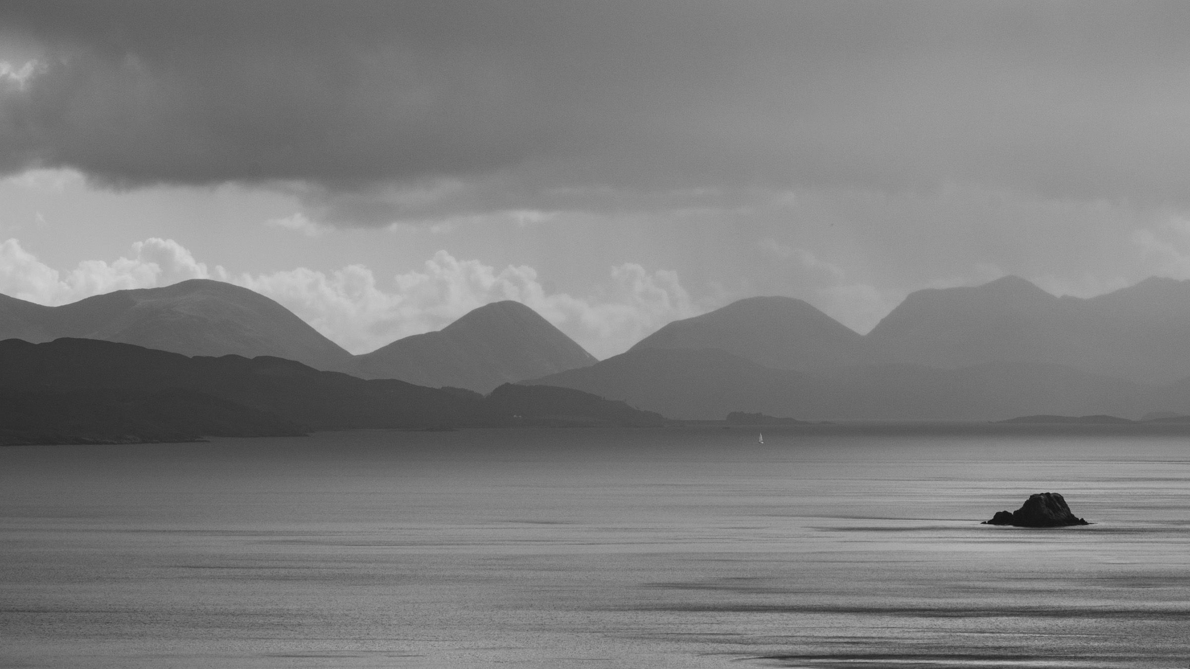 Black and white view of layered, rounded mountains across a calm sea, with a small rock in the water