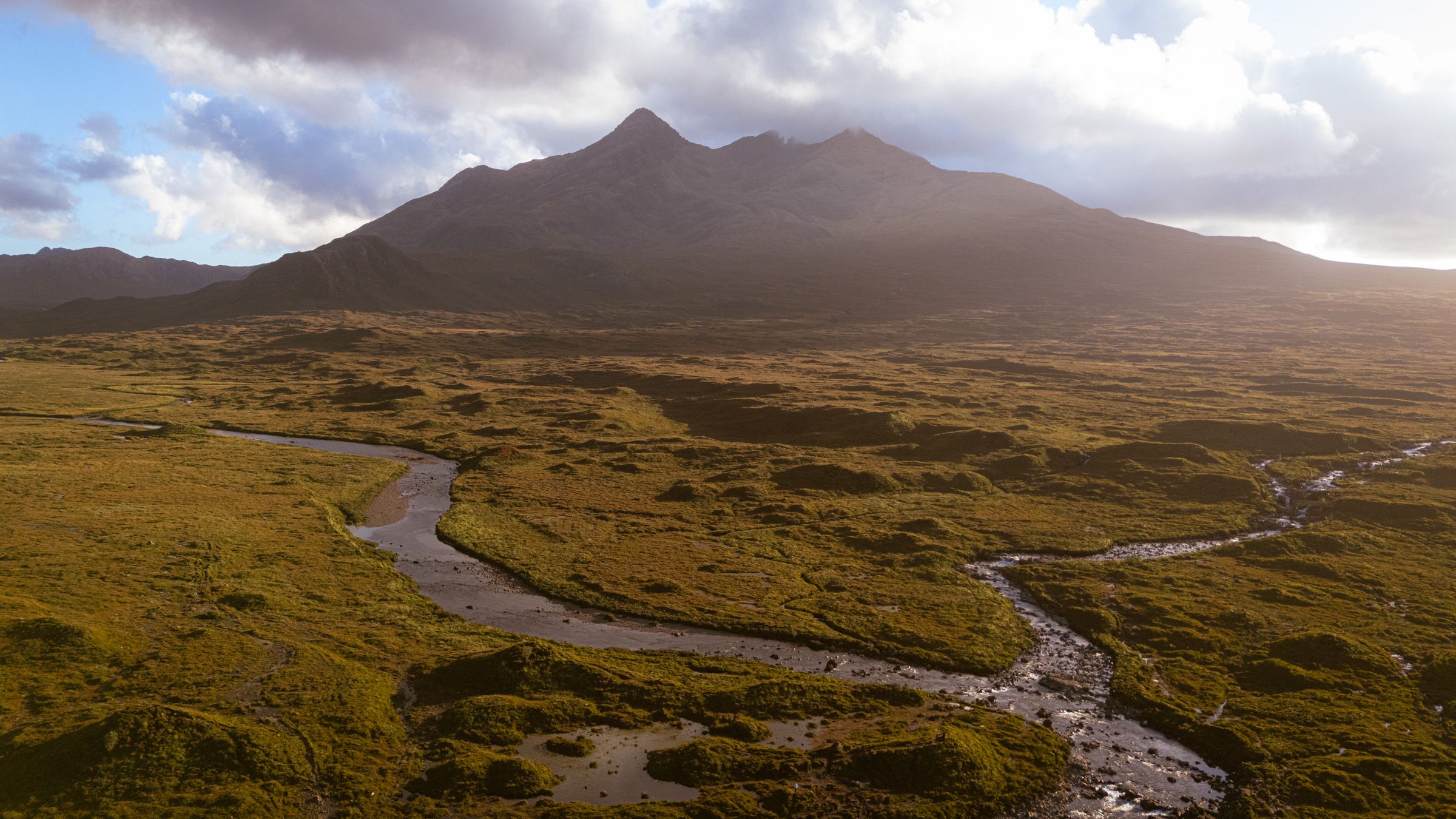 Aerial view of a river winding across golden moorland towards a dark, jagged mountain ridge under cloud