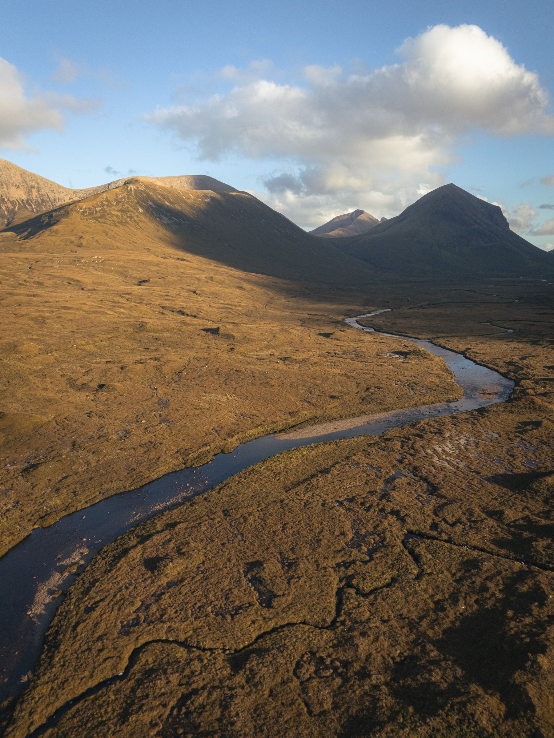Aerial view of a river curving through golden moorland towards sunlit hills, with a white cloud above