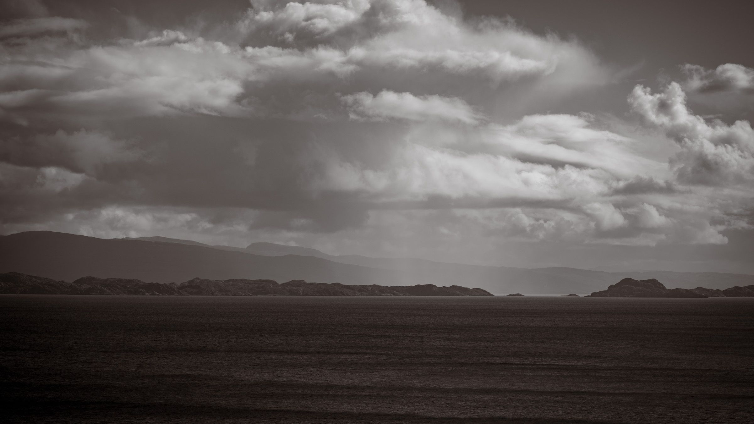 Toned black and white view across dark water to low rocky islands and distant hills under heavy cloud