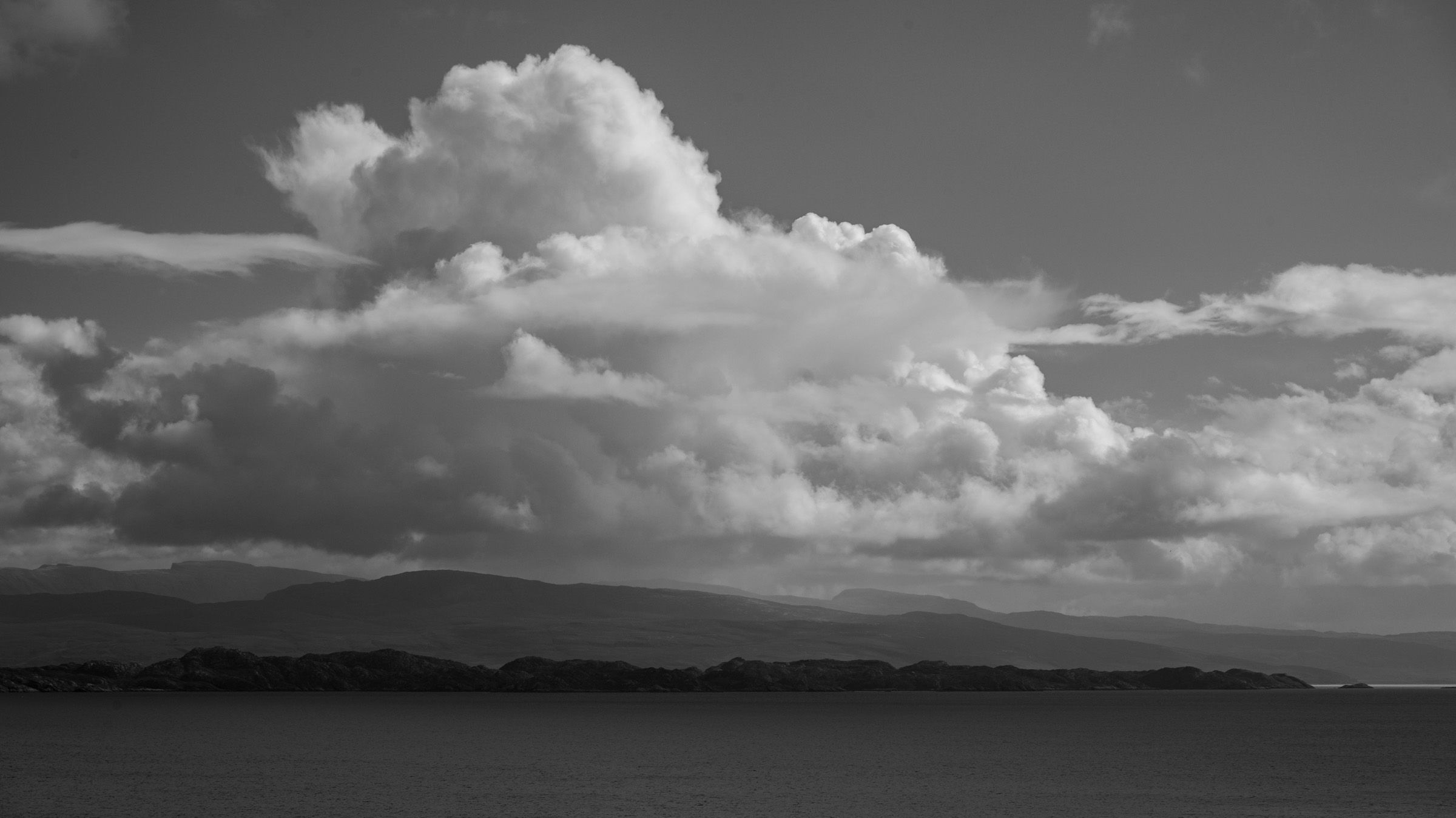 Black and white view of a large cumulus cloud above dark hills and still water