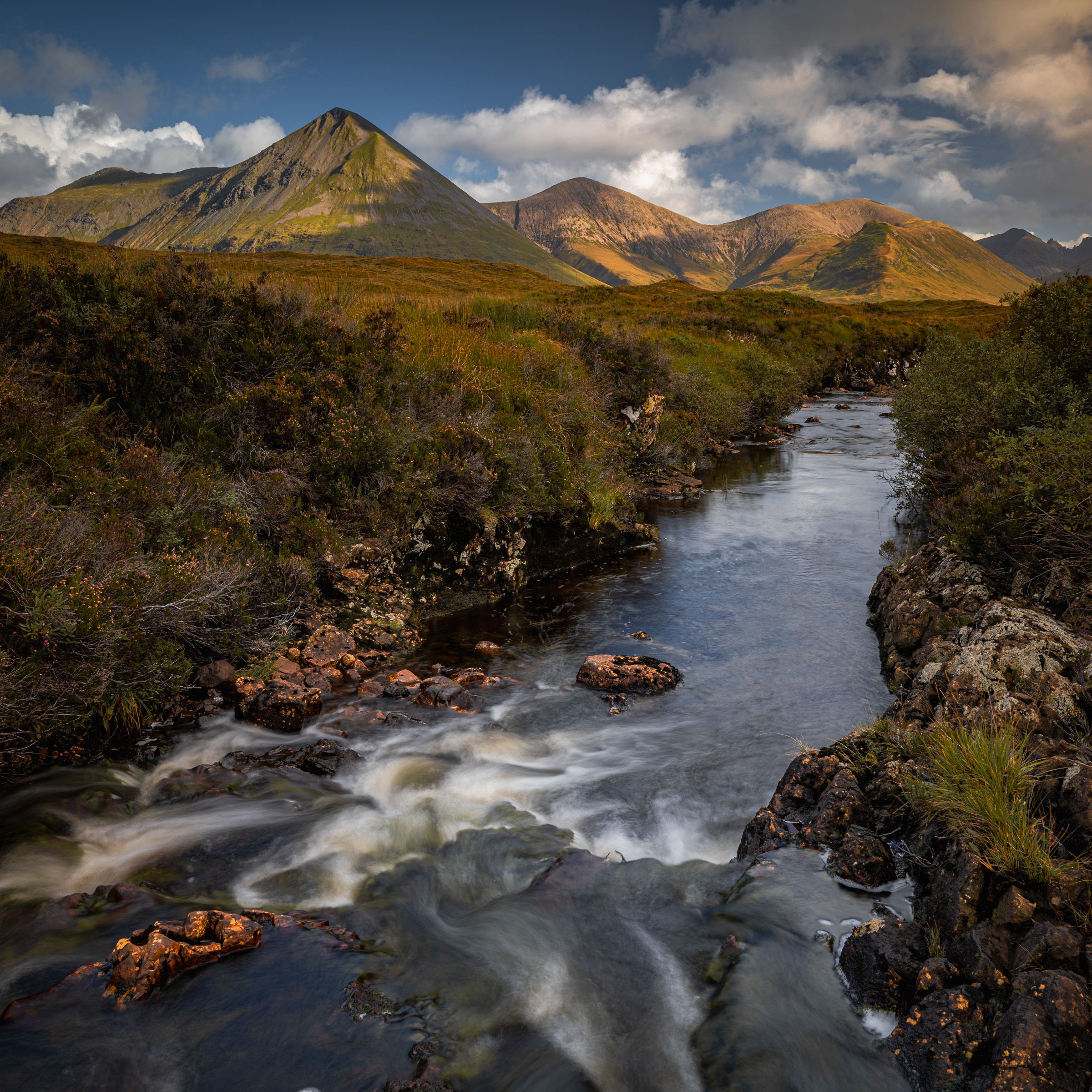 A stream cascading over rocks in the foreground, with a conical mountain and sunlit hills beyond